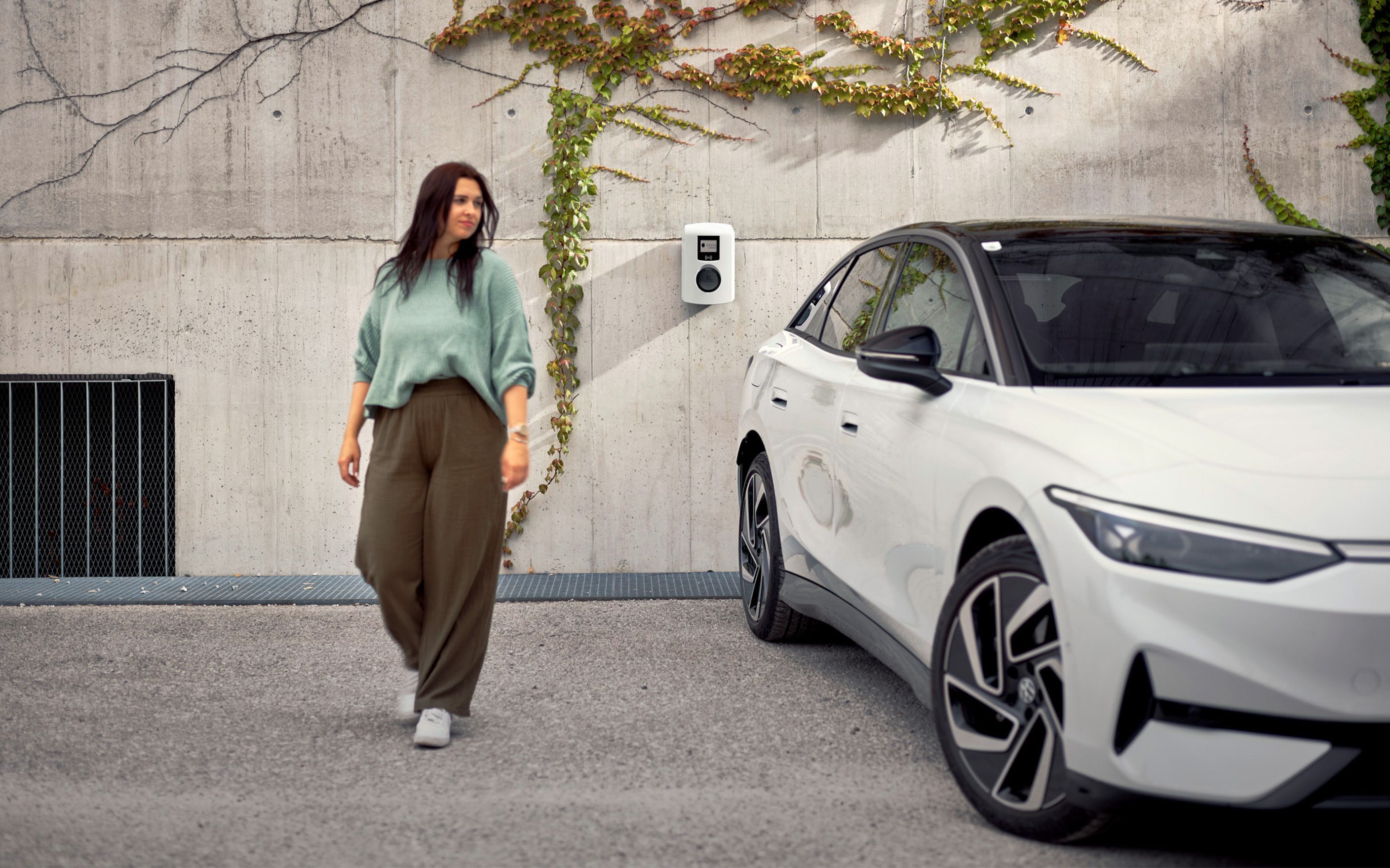 Жена върви до бял автомобил и зад нея стена с монтирана станция