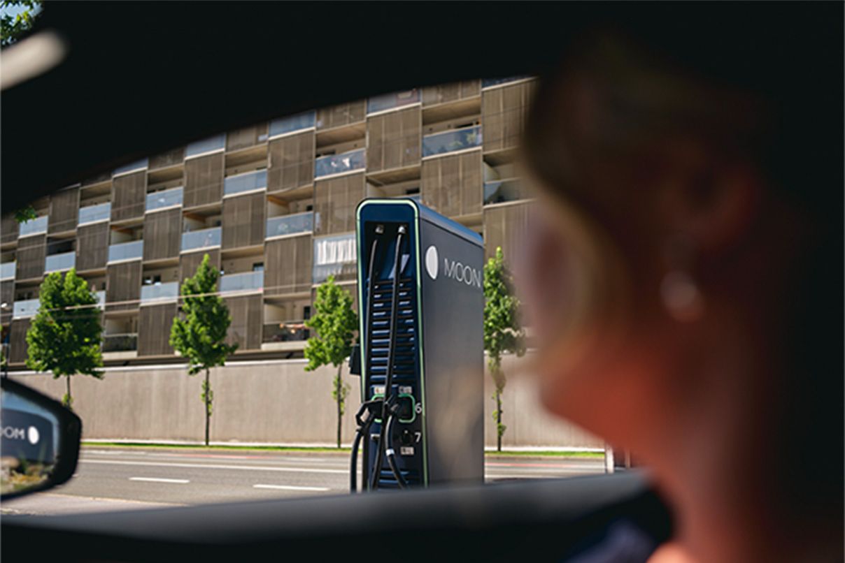 Човек, седнал в кола, а бърза зарядна станция се вижда от страничното стъкло