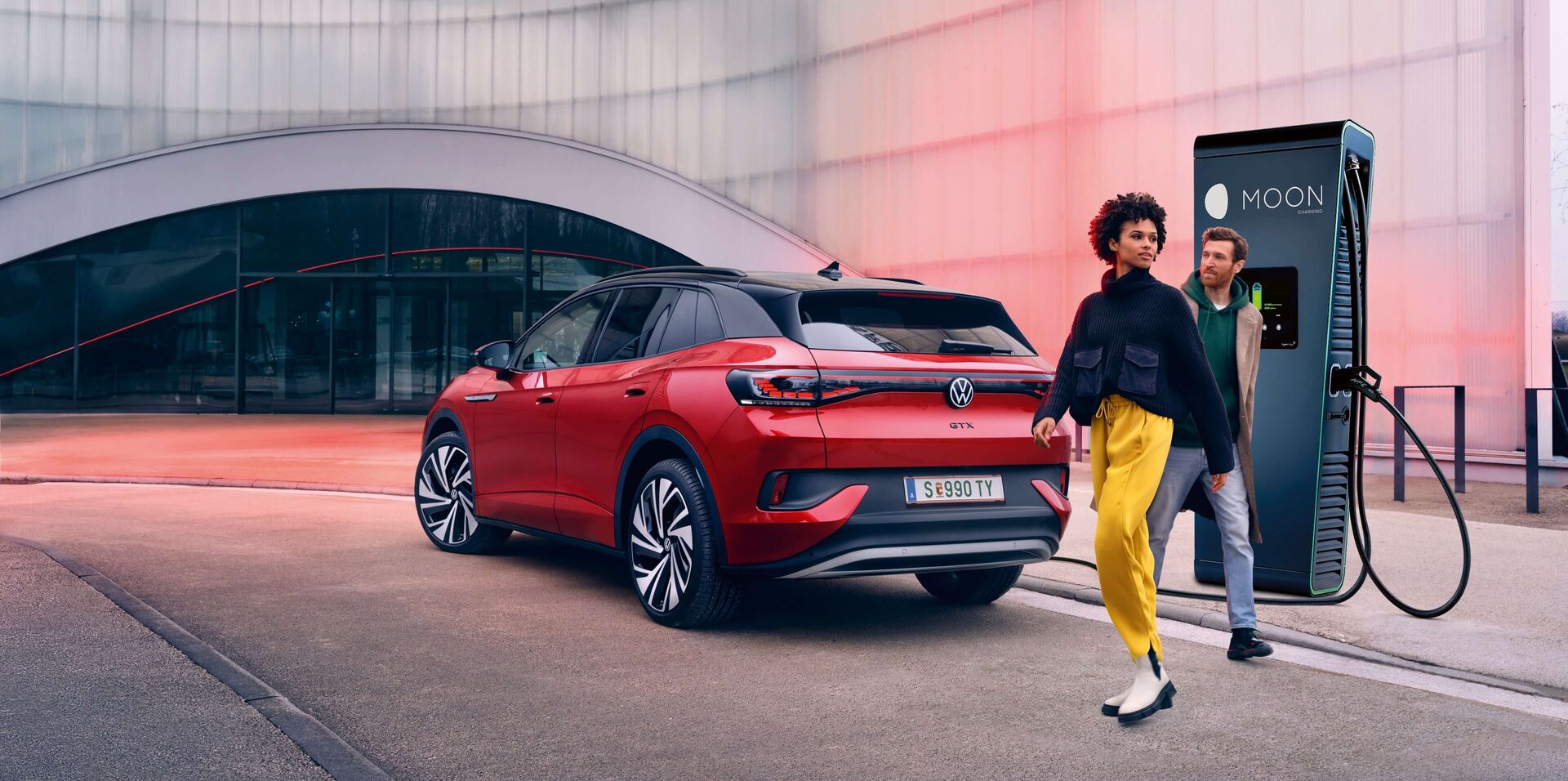 Спрял до хиперчарджър червен VW ID. автомобил. Зад него има жена с жълт панталон и черна блуза и мъж с дънки и половер