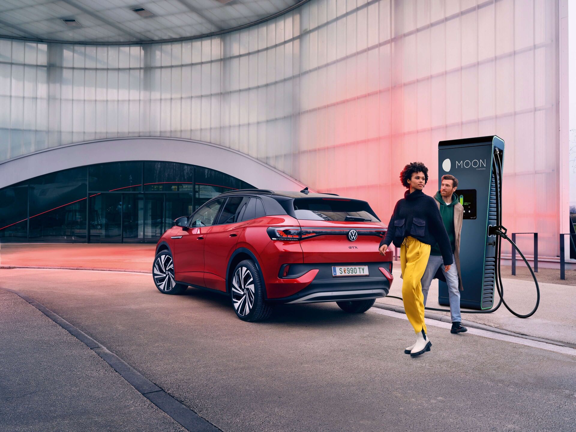 прял до хиперчарджър червен VW ID. автомобил. Зад него има жена с жълт панталон и черна блуза и мъж с дънки и половер