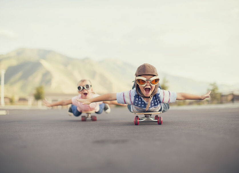Две деца, легнали върху скейборд дъска с каски и очила и разперени ръце.